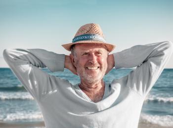 Een meneer op leeftijd zit te genieten bij de zee en vouwt zijn armen achter zijn hoofd