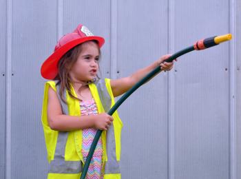 Klein meisje met helm en tuinslang speelt voor brandweervrouw